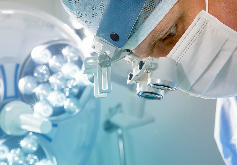 Close-up of an eye surgeon wearing surgical loupes and a mask during a cataract operation under bright operating room lights.